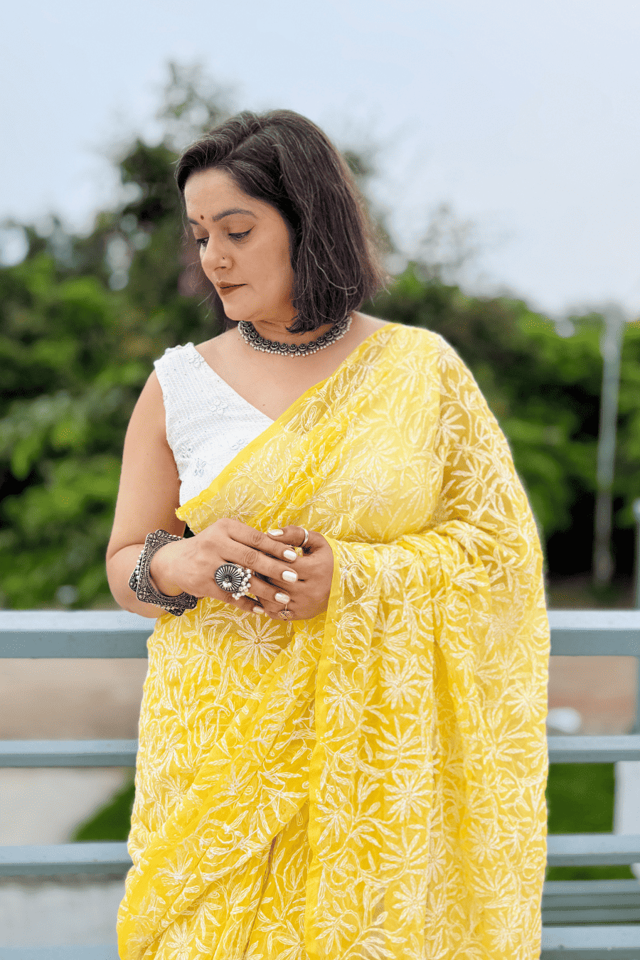 A woman wearing a bright yellow saree with white floral block prints and a white sleeveless blouse, standing outdoors with a blurred green background.