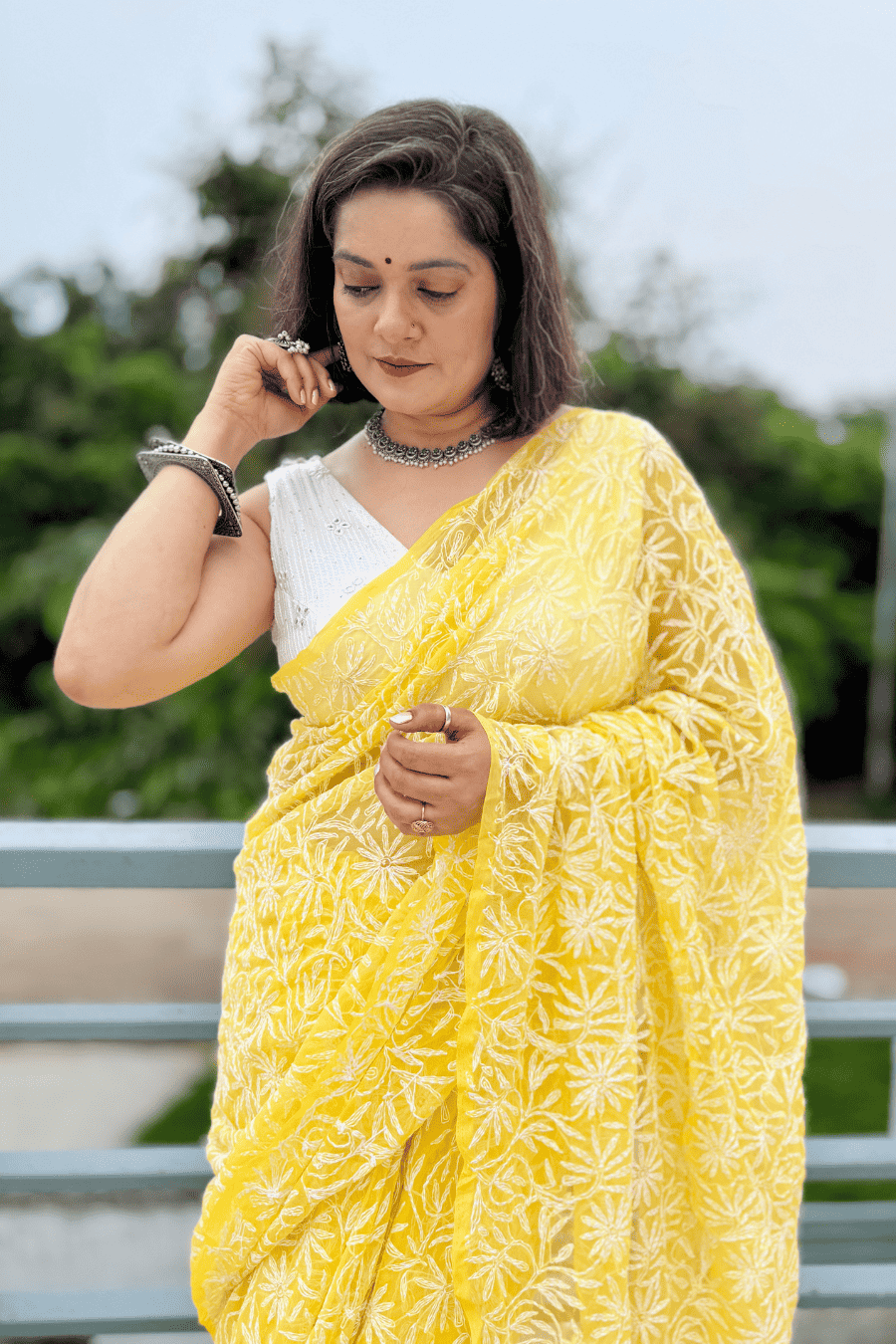 A woman wearing a bright yellow saree with white floral embroidery and a white blouse, standing outdoors with a blurred green background.