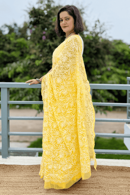 A woman wearing a bright yellow embroidered kurta with a matching dupatta, standing on a balcony with green foliage in the background.