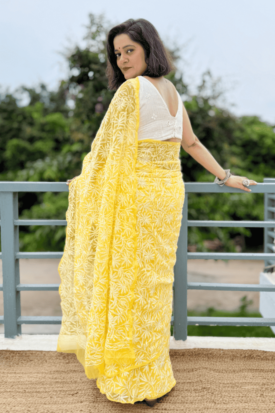 A woman wearing a bright yellow embroidered saree with a white blouse, standing on a balcony with a railing and green foliage in the background.