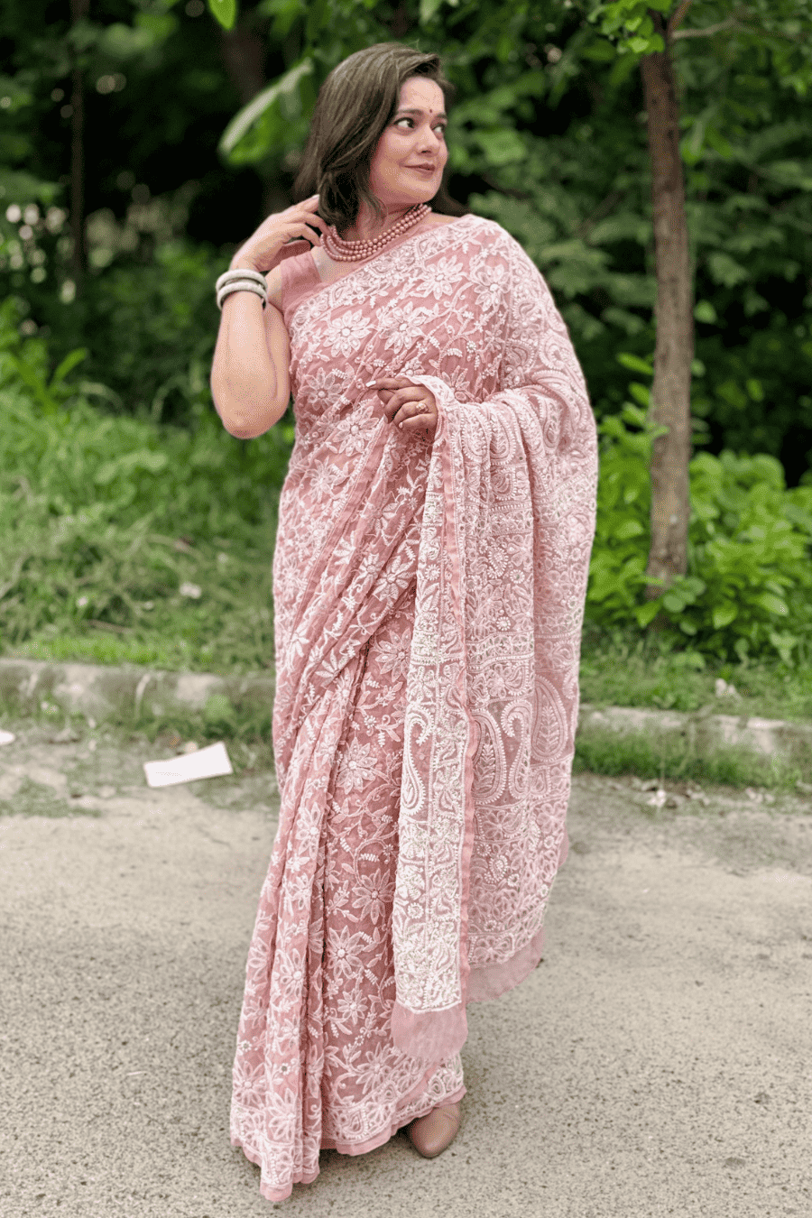 A woman wearing a pink and white floral lace saree stands outdoors on a paved path with green trees in the background.