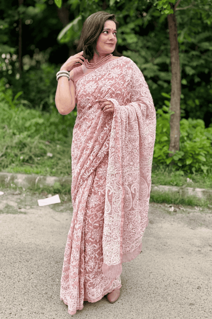 A woman wearing a pink and white floral lace saree stands outdoors on a paved path with green trees in the background.