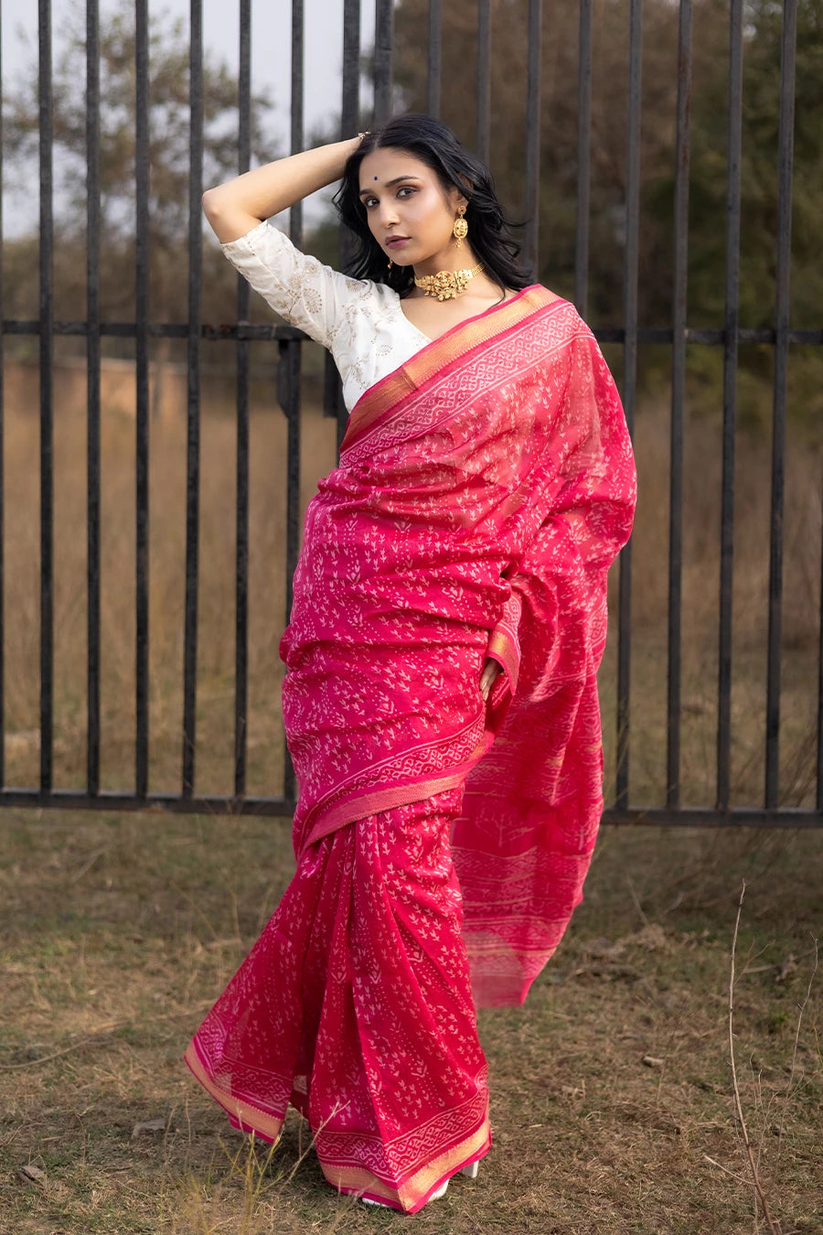 Woman in a red saree with white patterns standing outdoors near a metal fence.