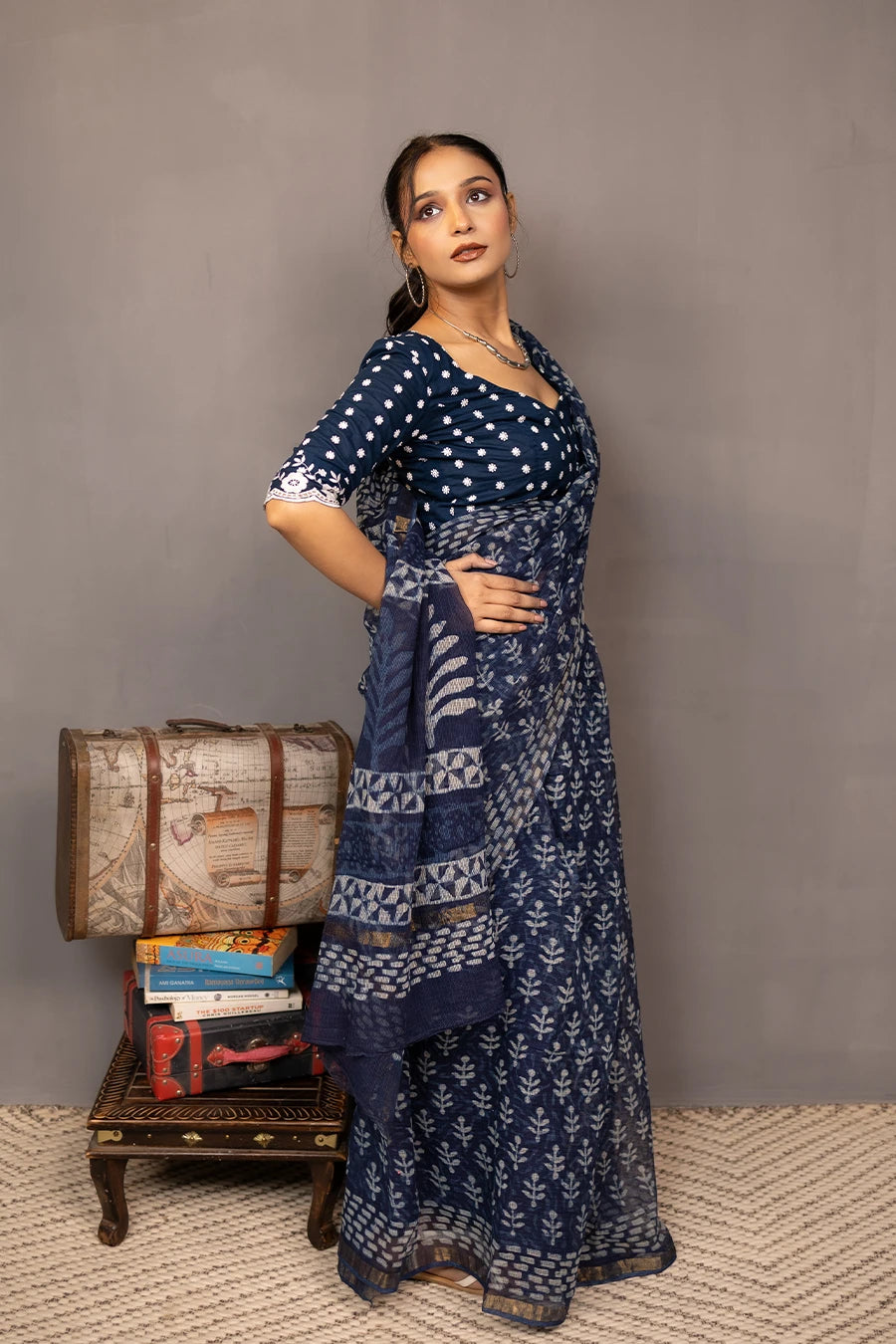 Woman in a patterned outfit standing next to a small table with books and a suitcase against a plain background