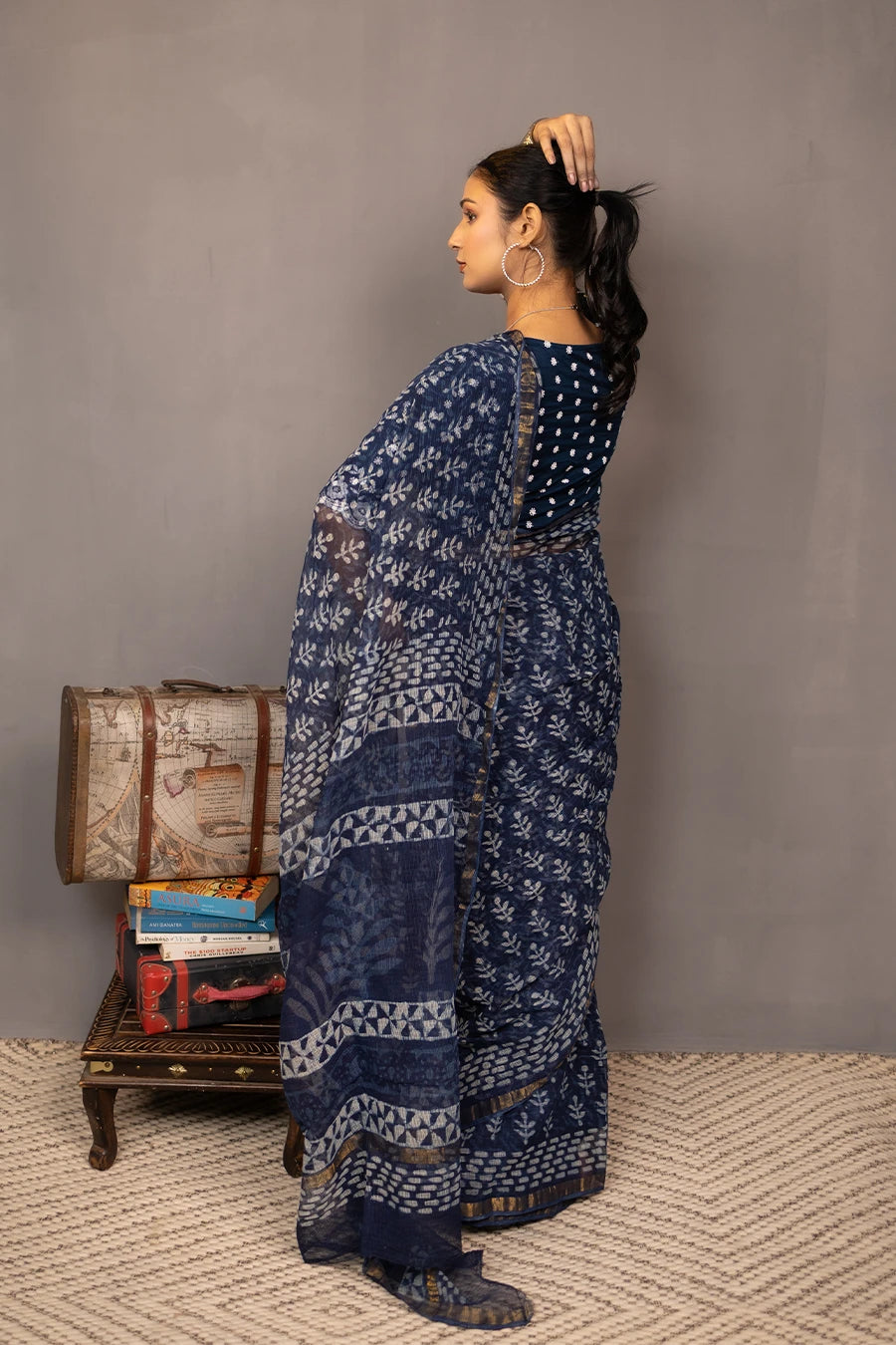 Woman in a patterned outfit standing next to a small table with books and a suitcase against a plain background