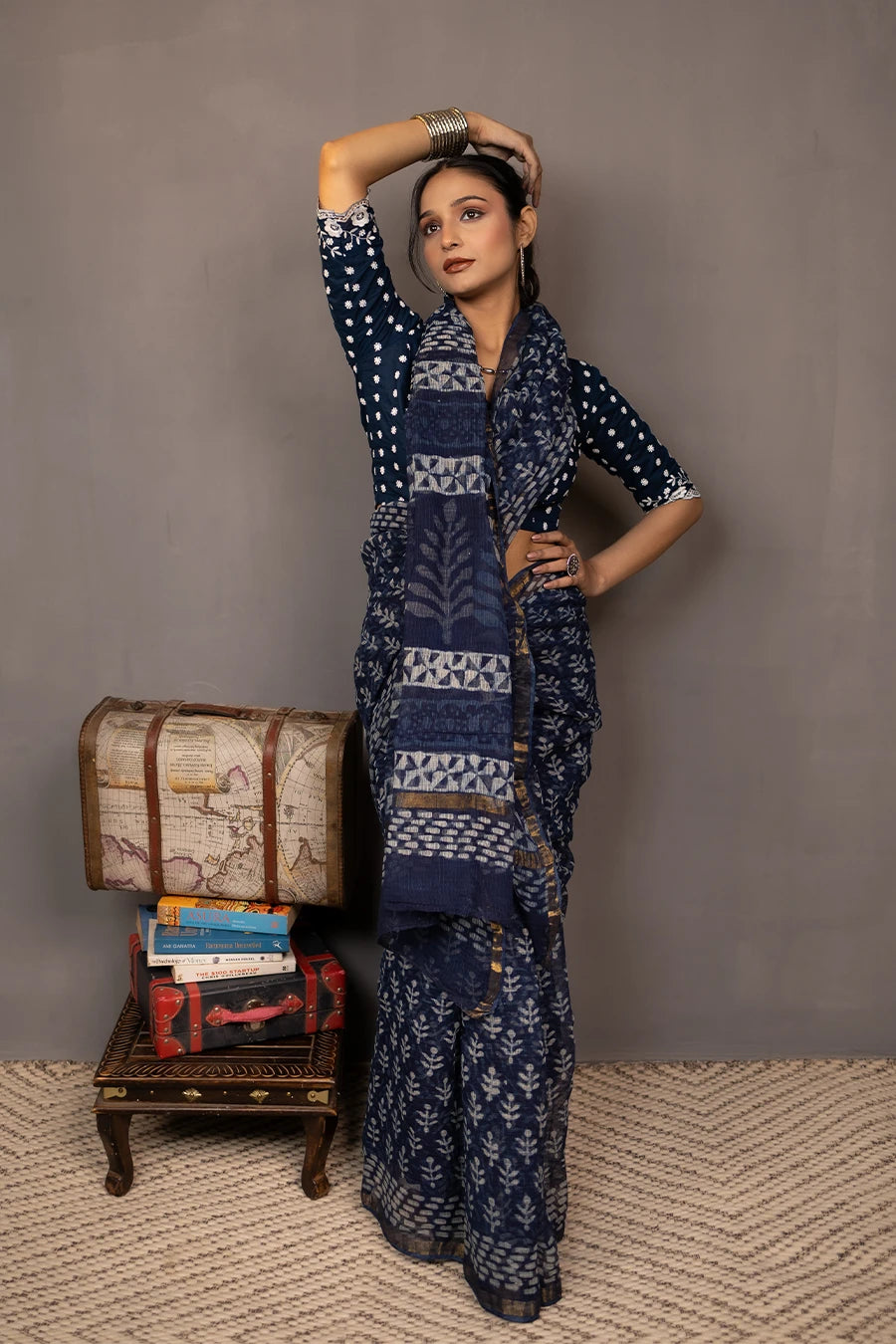 Woman in a patterned outfit standing next to a small table with books and a suitcase against a plain background