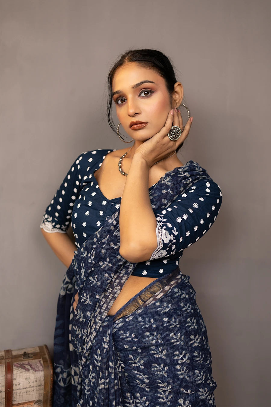 Woman in a patterned outfit standing next to a small table with books and a suitcase against a plain background