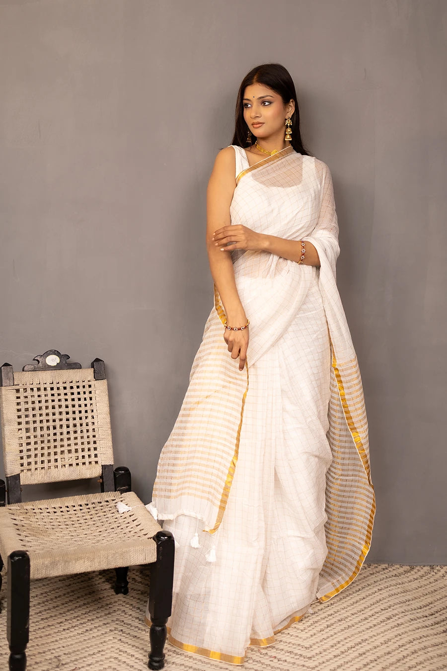 Woman in a white saree with gold border standing next to a wooden chair against a grey wall.