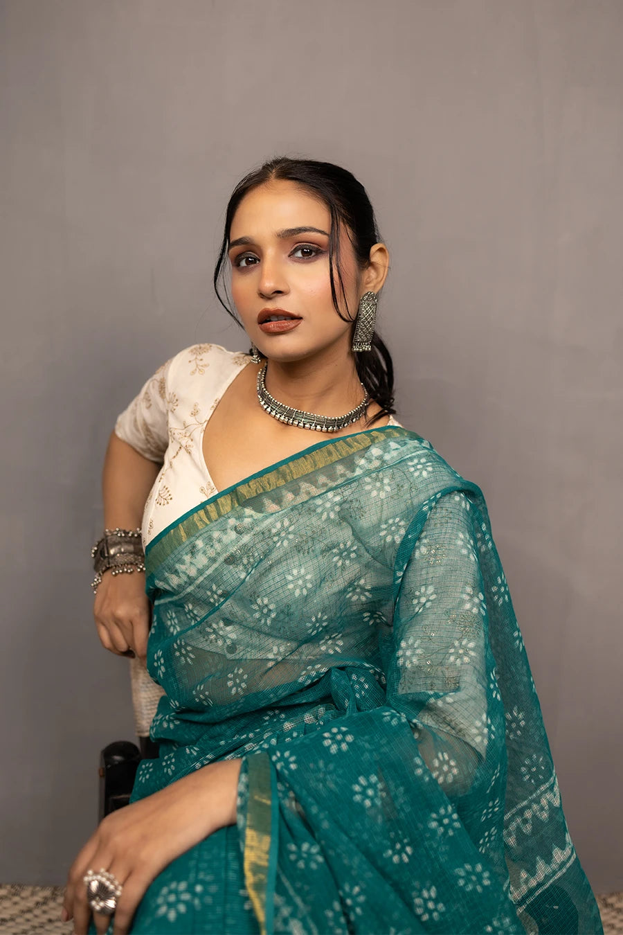 Woman in a teal saree standing next to a chair against a grey wall.