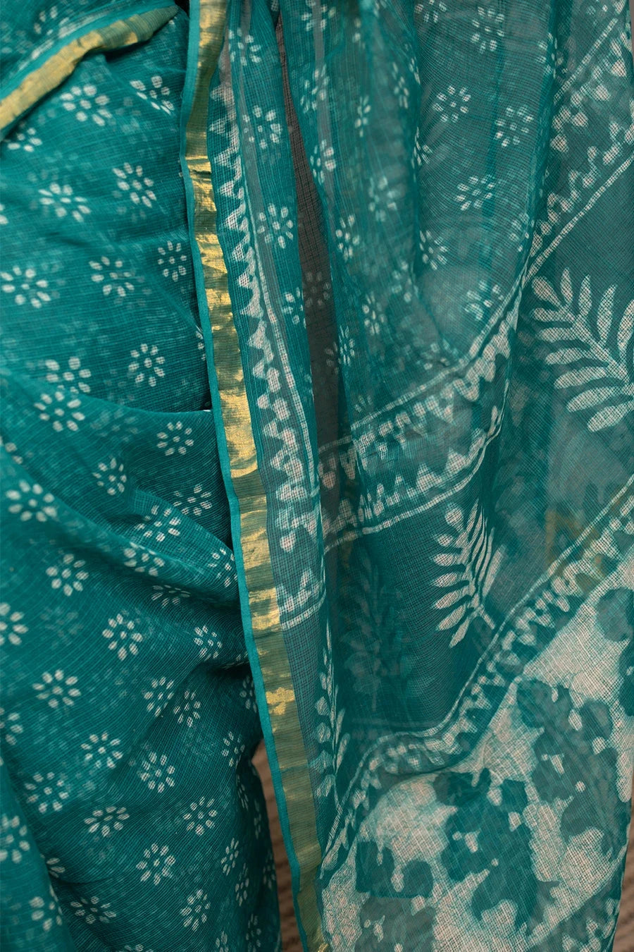 Woman in a teal saree standing next to a chair against a grey wall.