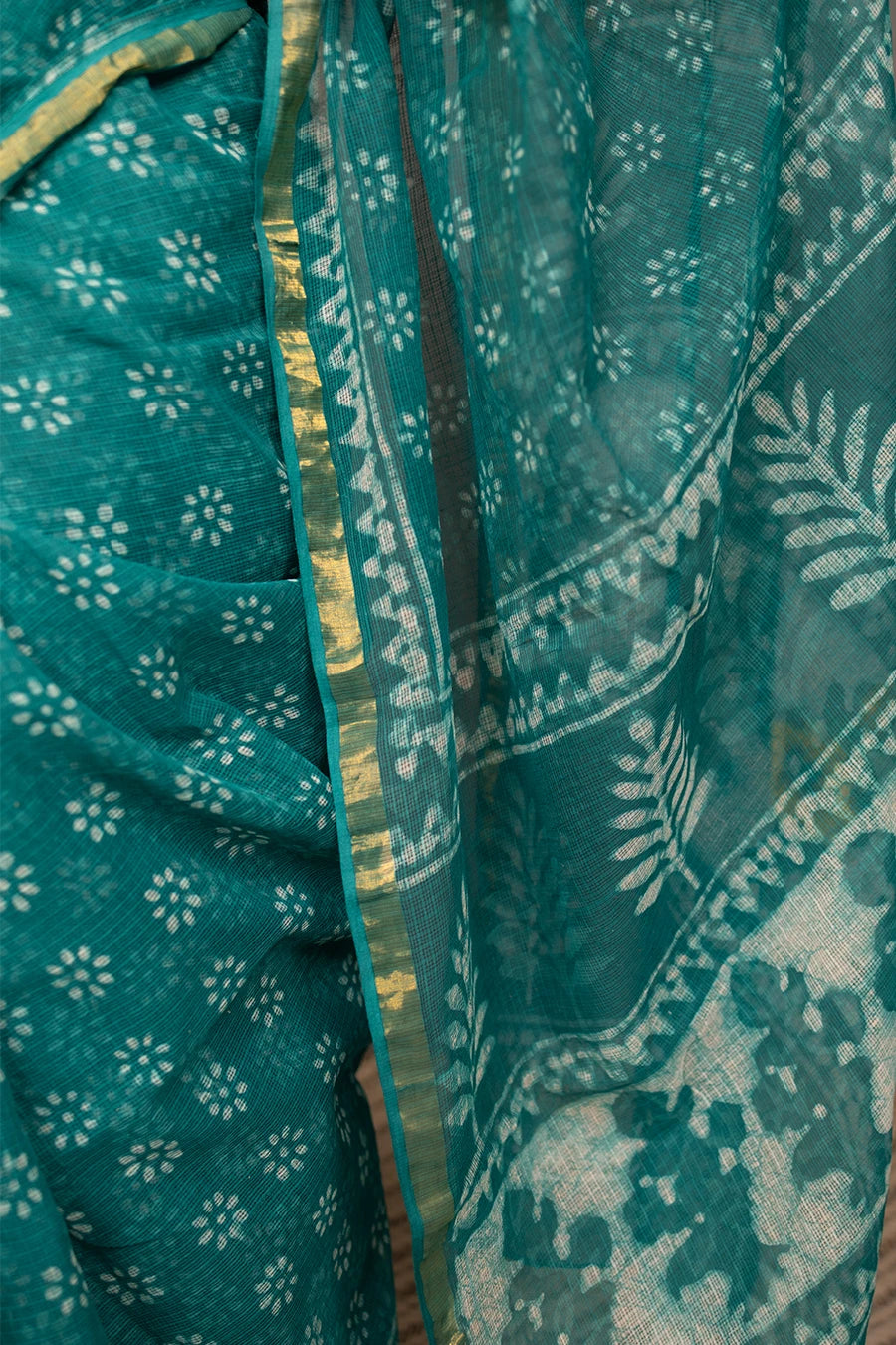 Woman in a teal saree standing next to a chair against a grey wall.