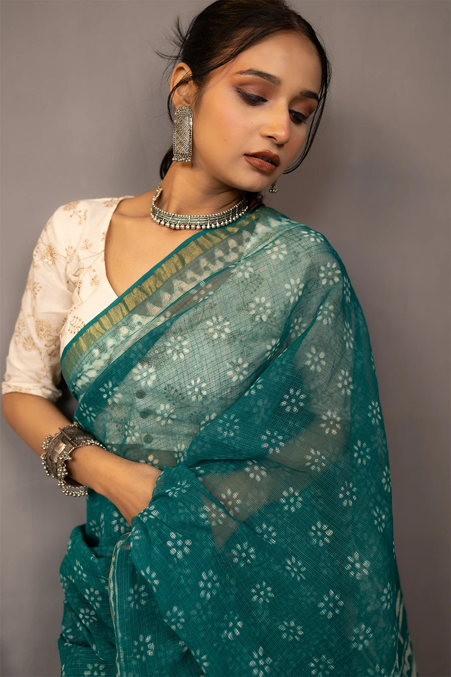 Woman in a teal saree standing next to a chair against a grey wall.