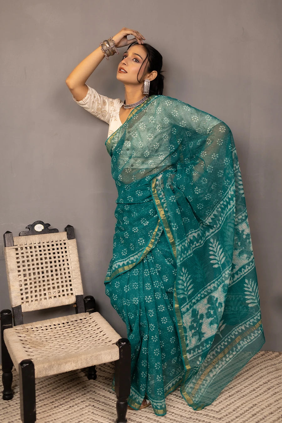 Woman in a teal saree standing next to a chair against a grey wall.