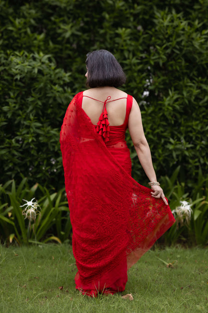 A woman wearing a bright red lace saree with a ruffled back, standing outdoors on grass in front of a dense green hedge.