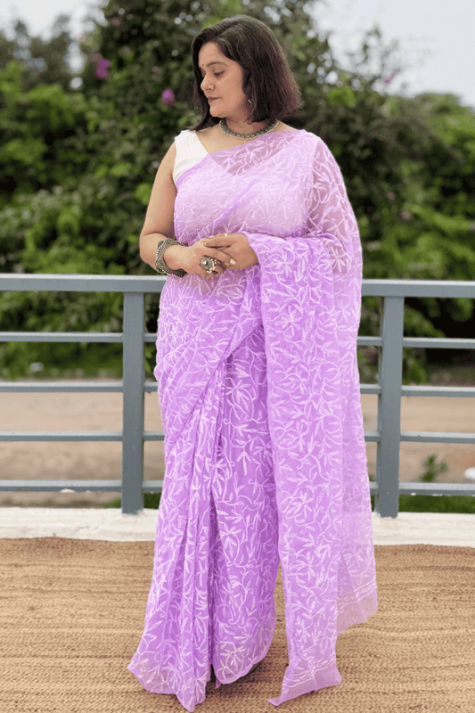 A woman wearing a lavender purple saree with white floral patterns and a white sleeveless blouse, standing outdoors in front of a railing with green foliage in the background.