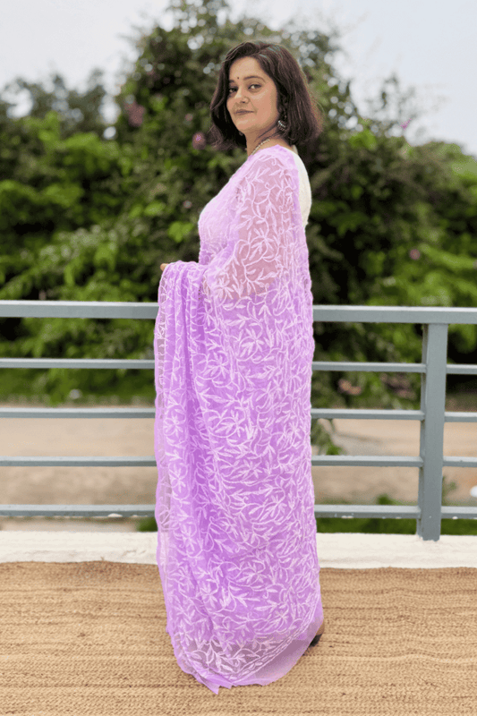 A woman wearing a lavender purple embroidered kurta and matching dupatta, standing outdoors on a jute mat with a metal railing and green foliage in the background.