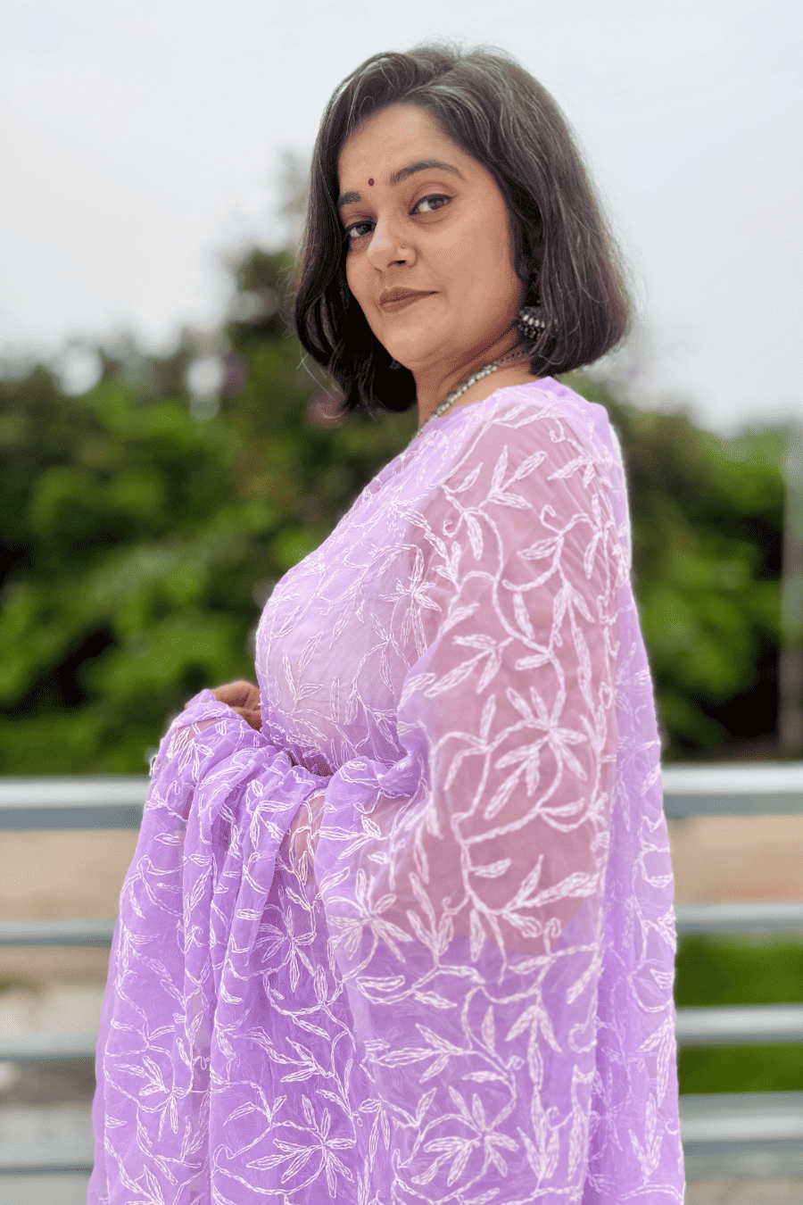 A woman wearing a lavender purple embroidered kurta with a matching sheer dupatta, photographed outdoors with a blurred green background.