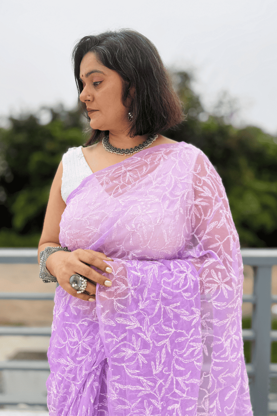A woman wearing a lavender purple embroidered saree with a white sleeveless blouse, standing outdoors with a blurred green background.