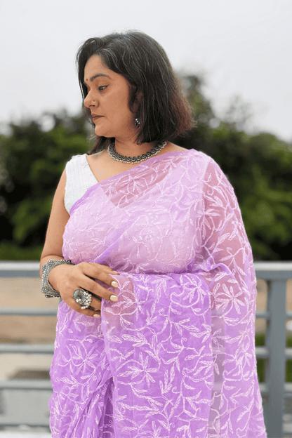 A woman wearing a lavender purple embroidered saree with a white sleeveless blouse, standing outdoors with a blurred green background.