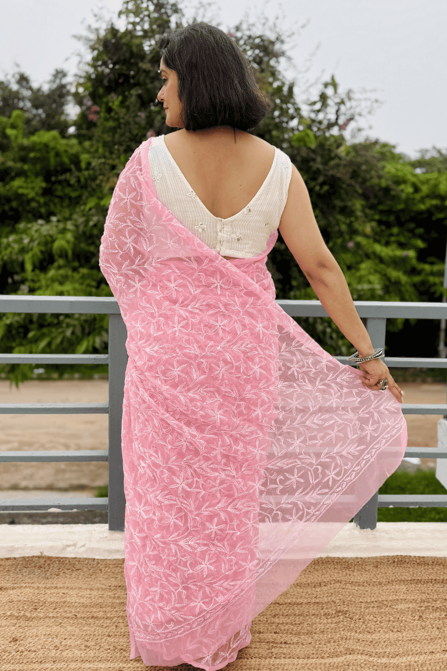 A woman wearing a light pink saree with white floral embroidery and a white sleeveless blouse, standing outdoors on a balcony with green foliage in the background.