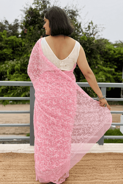 A woman wearing a light pink saree with white floral embroidery and a white sleeveless blouse, standing outdoors on a balcony with green foliage in the background.