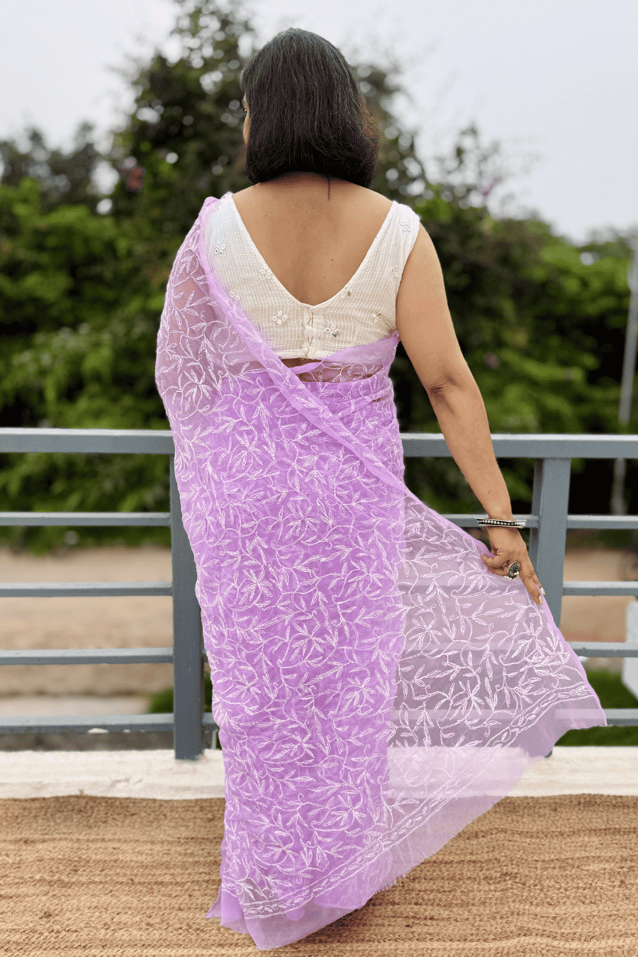 A woman shown from behind wearing a lavender purple embroidered saree with a white sleeveless blouse, standing outdoors on a balcony with green trees in the background.
