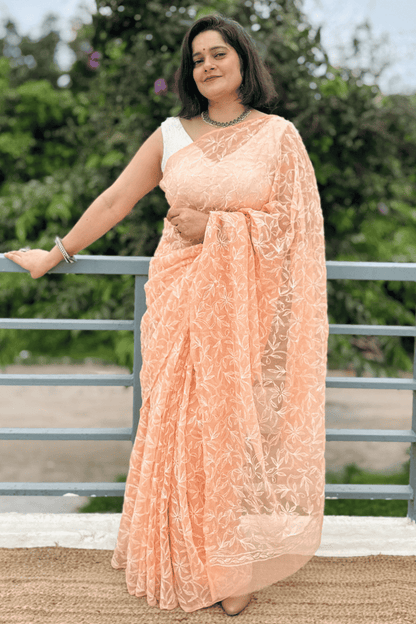 A woman wearing a peach-colored embroidered saree with a white sleeveless blouse, standing outdoors on a balcony with green foliage in the background.