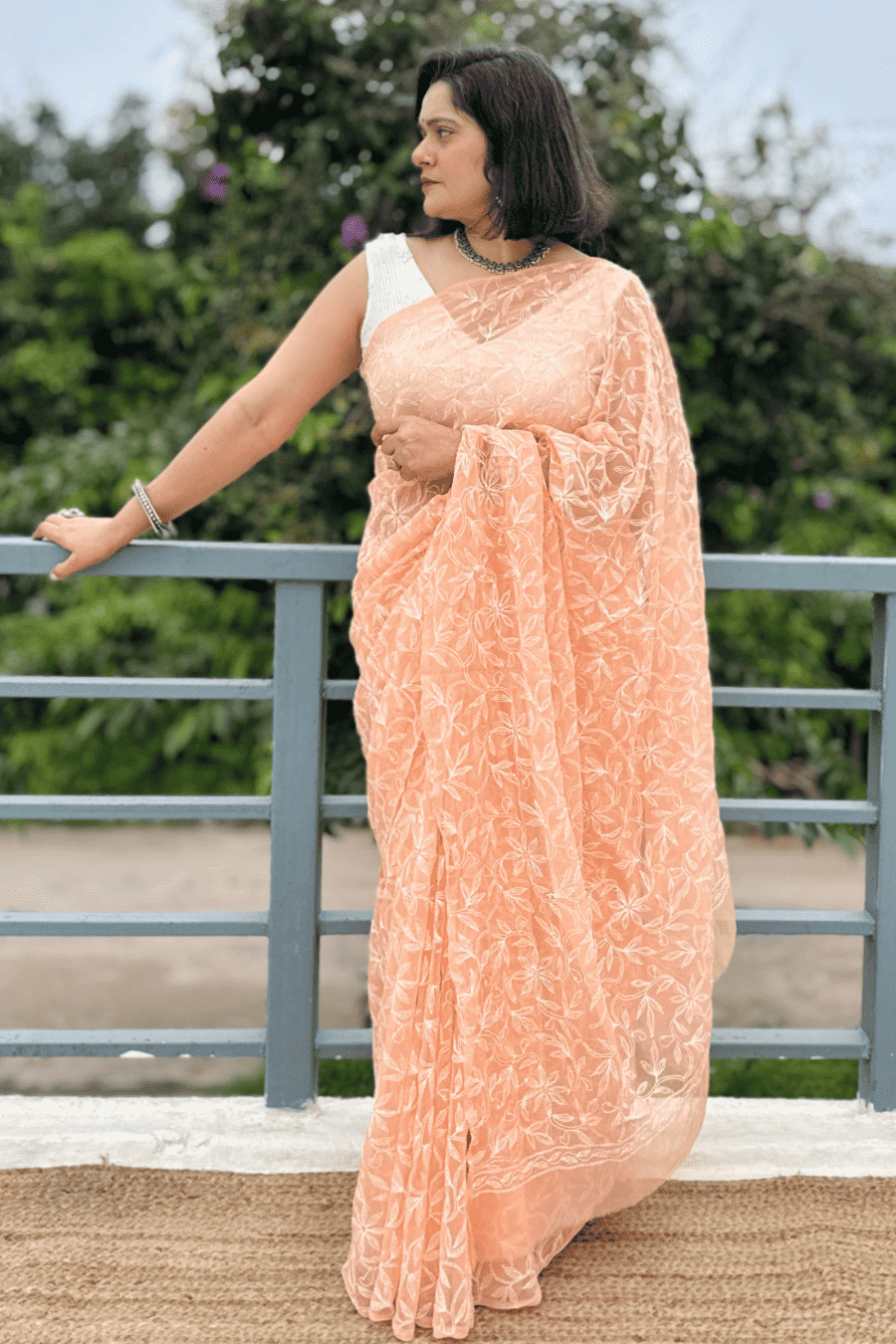 A woman wearing a peach-colored embroidered saree with a white sleeveless blouse, standing on a balcony with green foliage in the background.