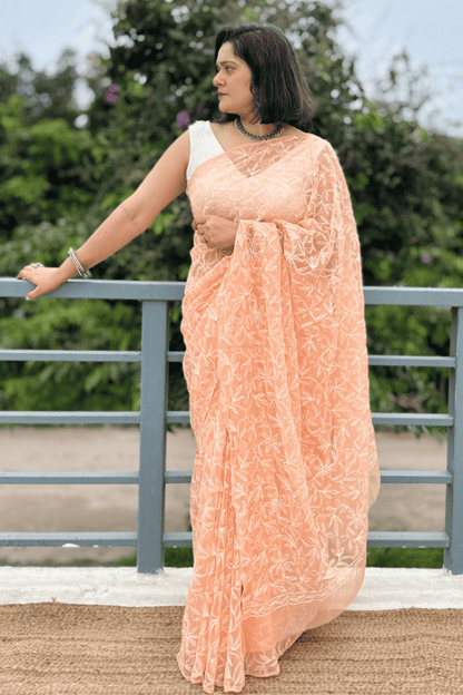 A woman wearing a peach-colored embroidered saree with a white sleeveless blouse, standing on a balcony with green foliage in the background.