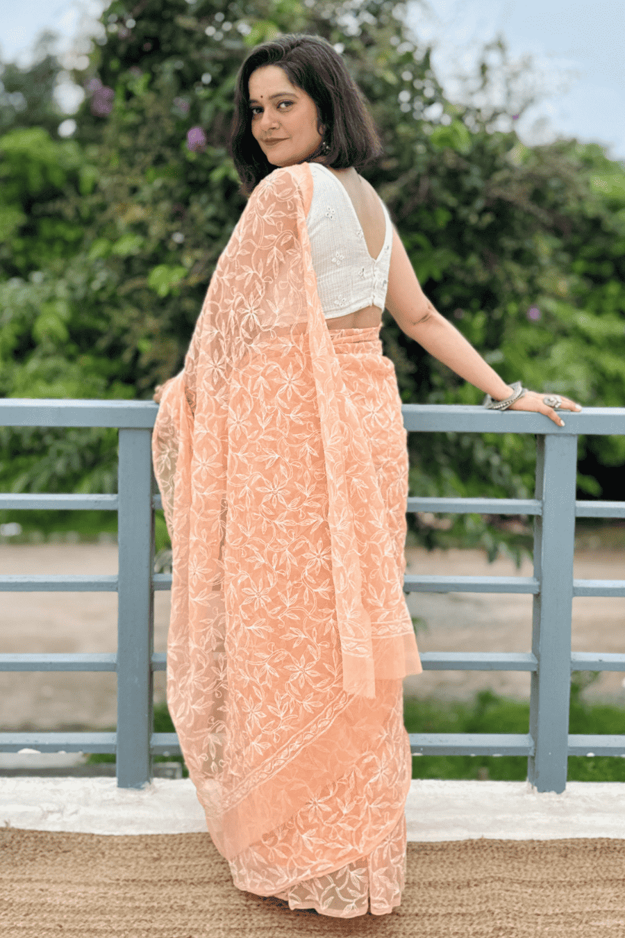 A woman wearing a peach-colored embroidered saree with a white sleeveless blouse, standing outdoors next to a gray railing with green foliage in the background.