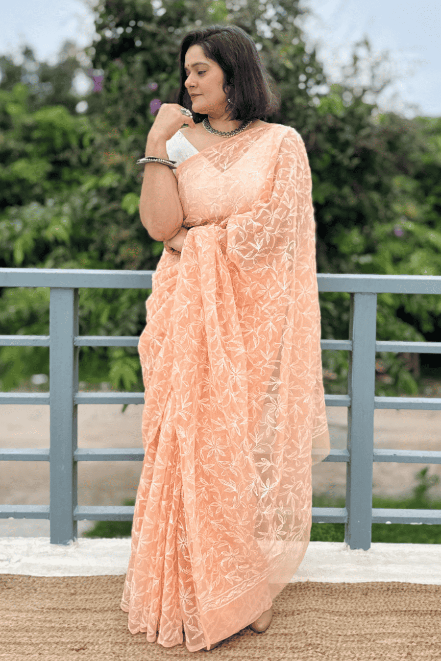 A woman wearing a peach-colored embroidered saree with a matching blouse, standing outdoors in front of a gray railing and green foliage.