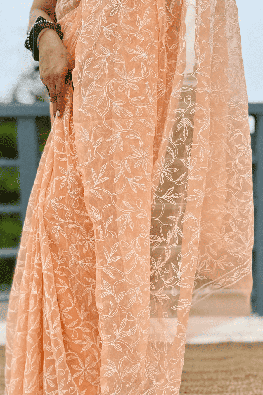 A close-up of a peach-colored embroidered saree or dress with white floral lace patterns, worn by a model whose hand and arm are visible.