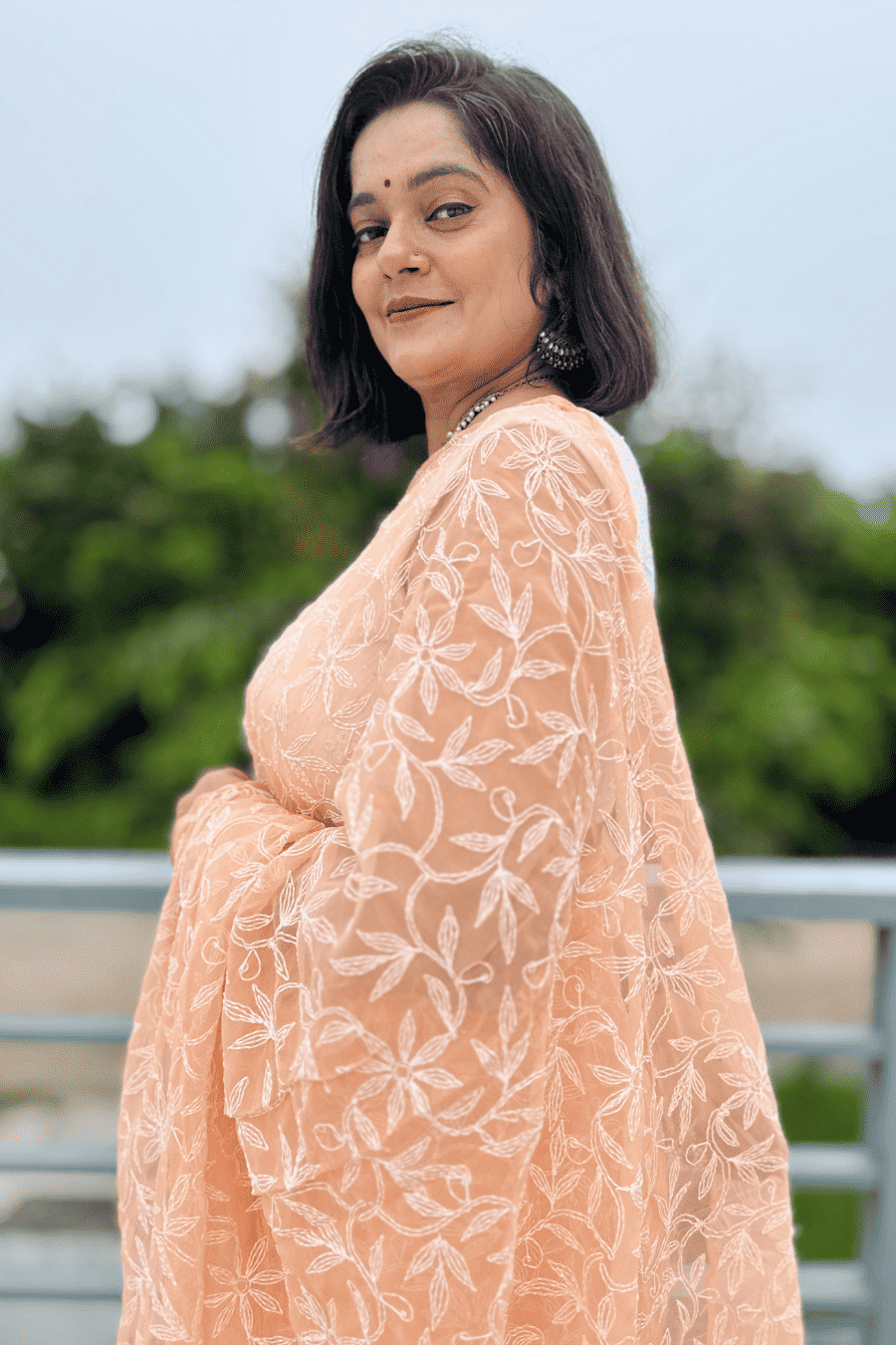 A woman wearing a peach-colored embroidered saree with white floral patterns, photographed outdoors with a blurred green background.