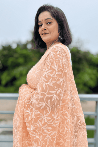 A woman wearing a peach-colored embroidered saree with white floral patterns, photographed outdoors with a blurred green background.