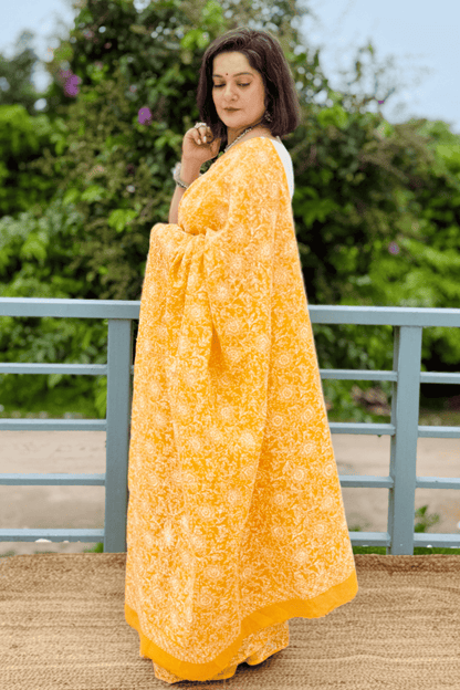 A woman wearing a bright yellow saree with white floral block prints stands outdoors on a balcony with green foliage in the background.