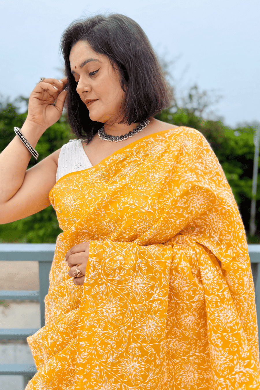 A woman wearing a bright yellow saree with a white floral block print pattern and a white sleeveless blouse, standing outdoors.