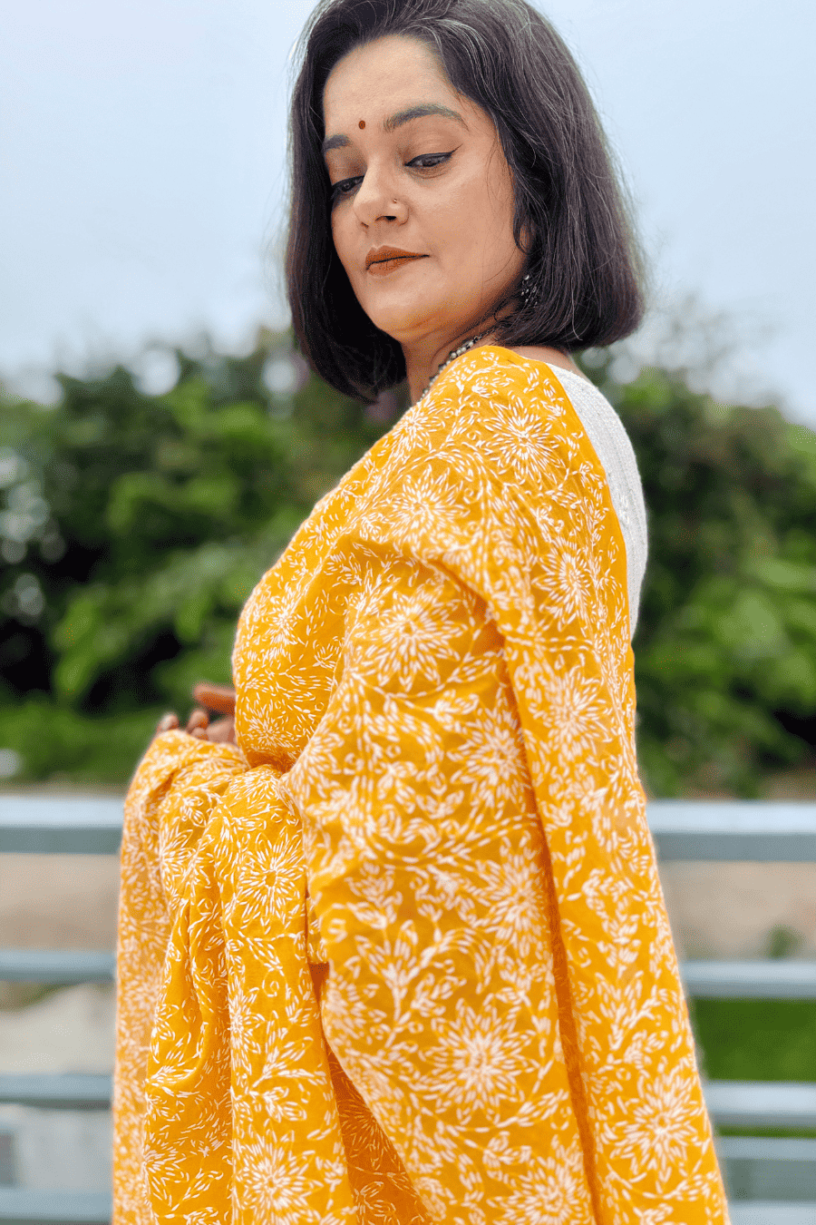 A woman wearing a bright yellow and white floral print kurta or sari, photographed outdoors with a blurred green background.