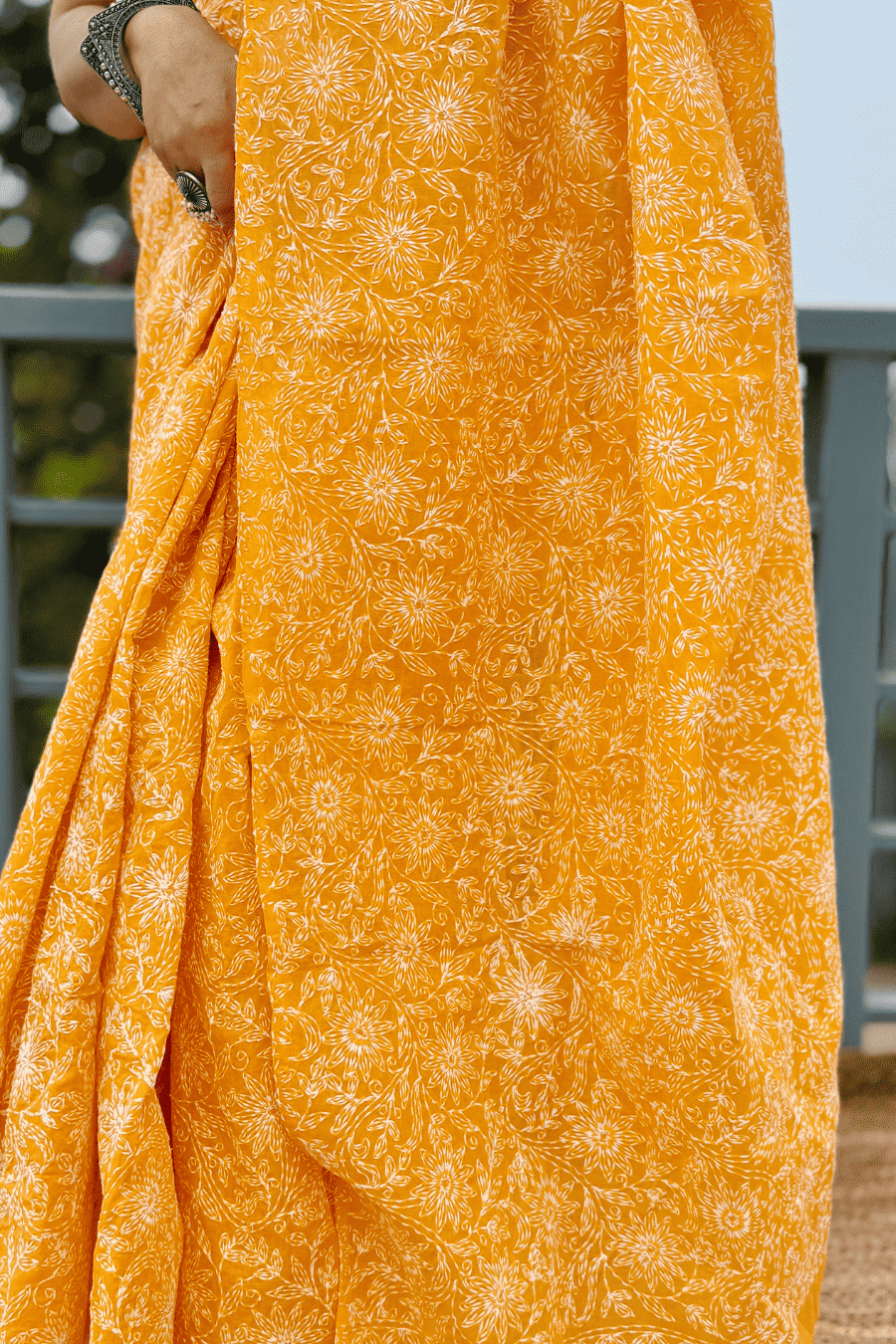 A close-up of a mustard yellow fabric saree with a white block-printed floral pattern, held by a person wearing a silver bracelet.