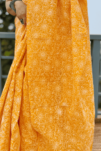 A close-up of a mustard yellow fabric saree with a white block-printed floral pattern, held by a person wearing a silver bracelet.