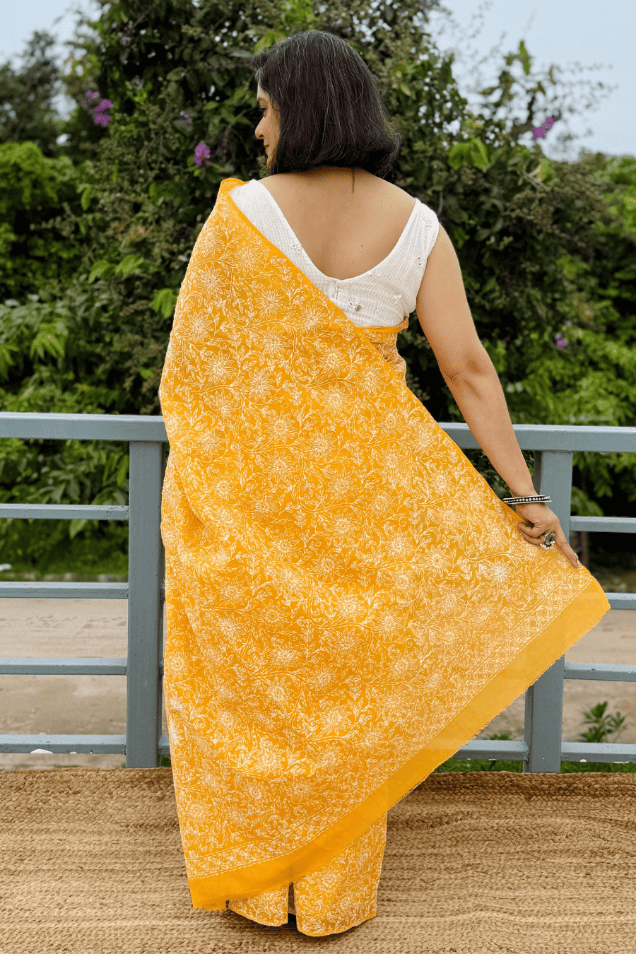 A woman wearing a bright yellow printed saree with a white sleeveless blouse, standing outdoors on a balcony with green foliage in the background.
