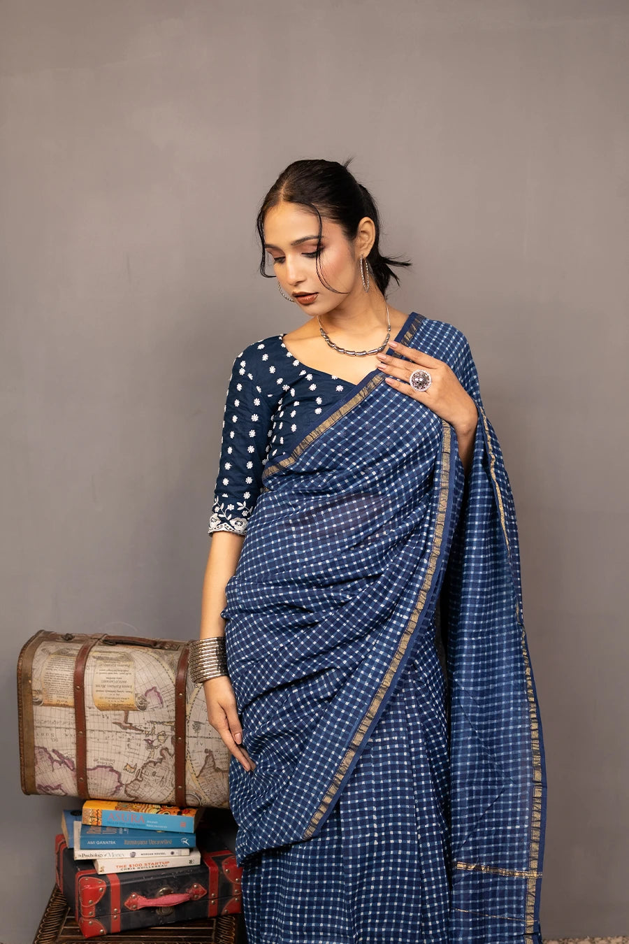 Woman in a blue saree standing next to a small wooden table with books and a suitcase against a grey background.