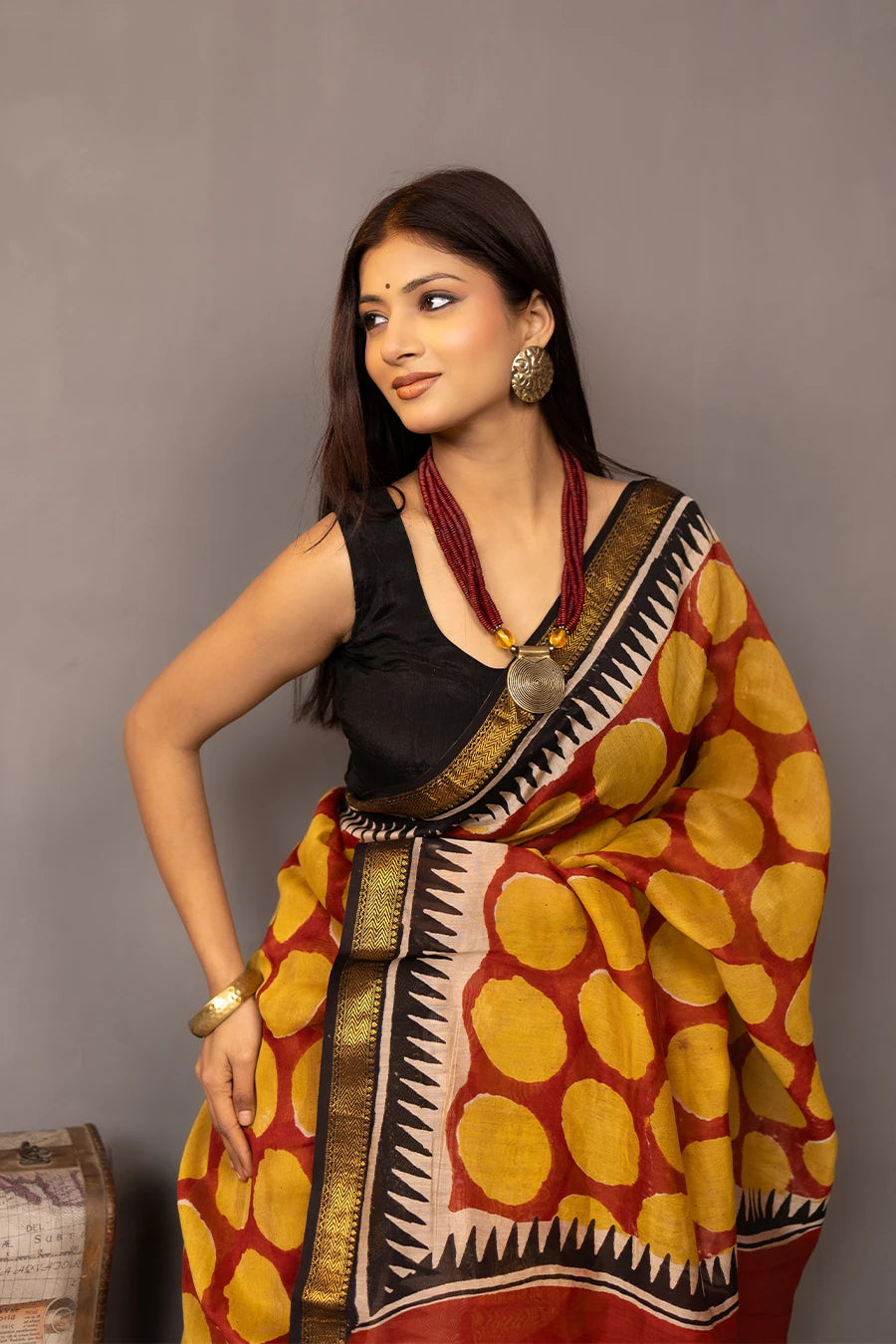 Woman wearing a traditional saree with a patterned design, standing next to a small table with books and a suitcase.
