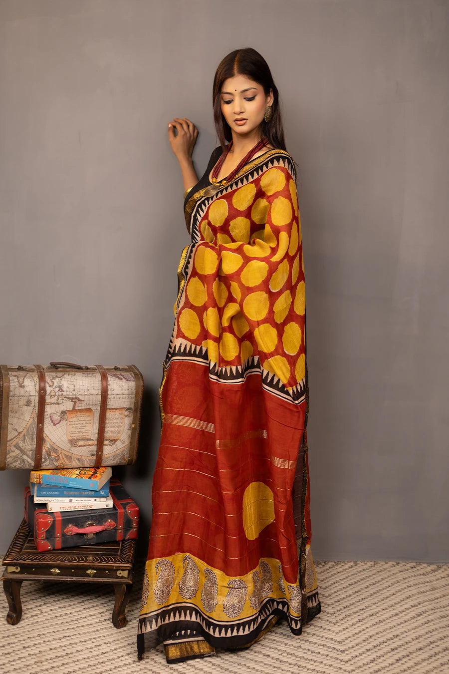 Woman wearing a traditional saree with a patterned design, standing next to a small table with books and a suitcase.