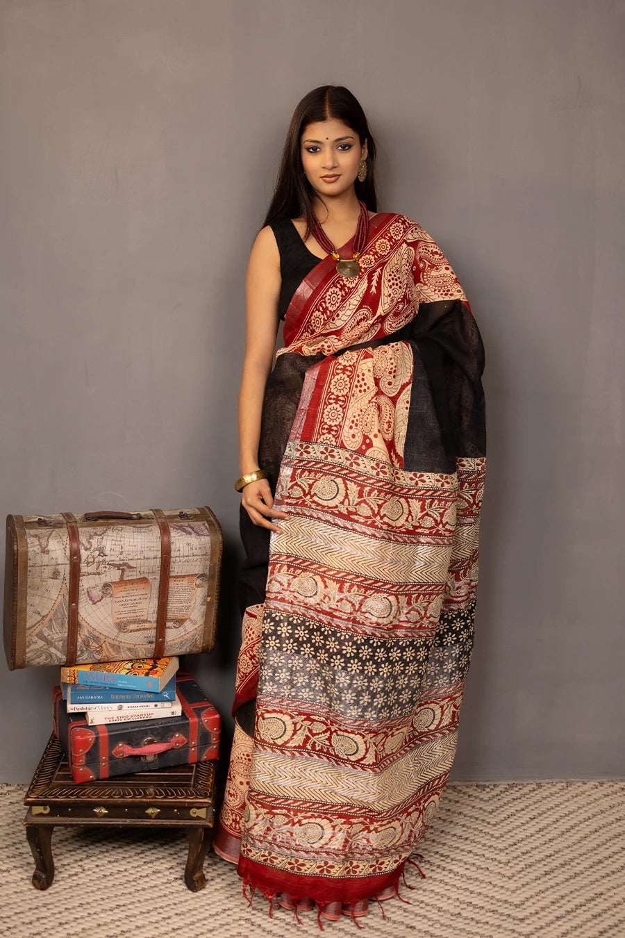 Woman wearing a traditional saree with a patterned design, standing next to a small table with books and a suitcase.