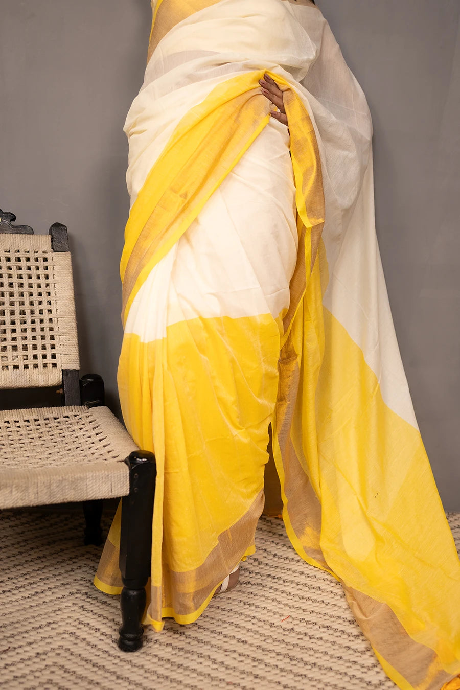 Woman in a yellow and white saree standing next to a chair against a grey background