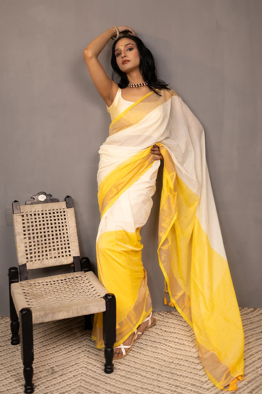Woman in a yellow and white saree standing next to a chair against a grey background