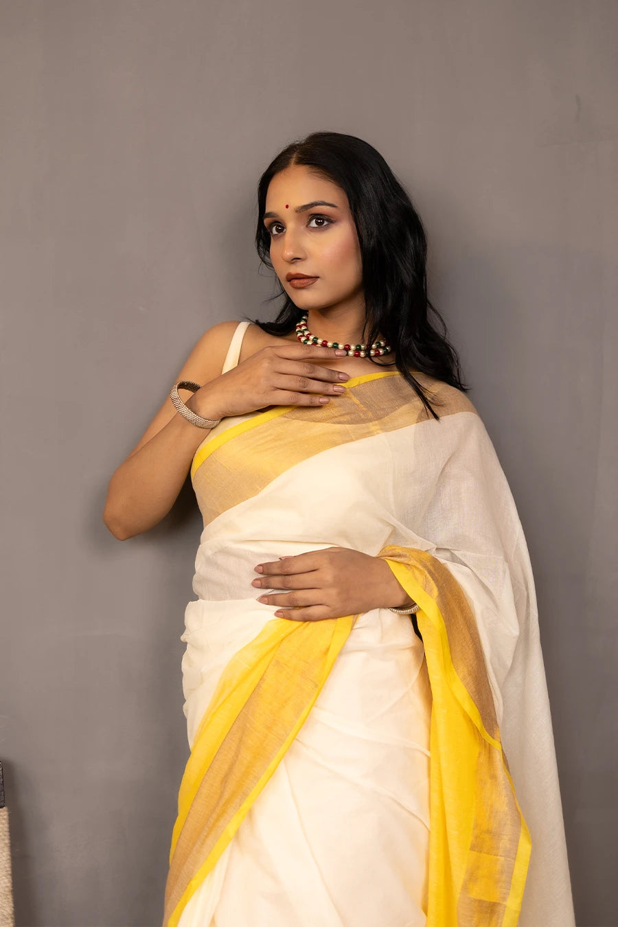 Woman in a yellow and white saree standing next to a chair against a grey background