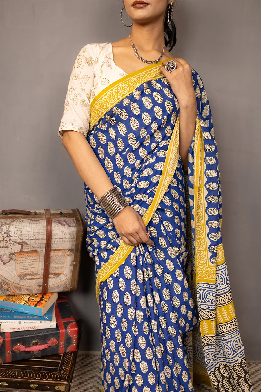 Woman wearing a blue and yellow saree standing next to vintage suitcases and books against a grey background
