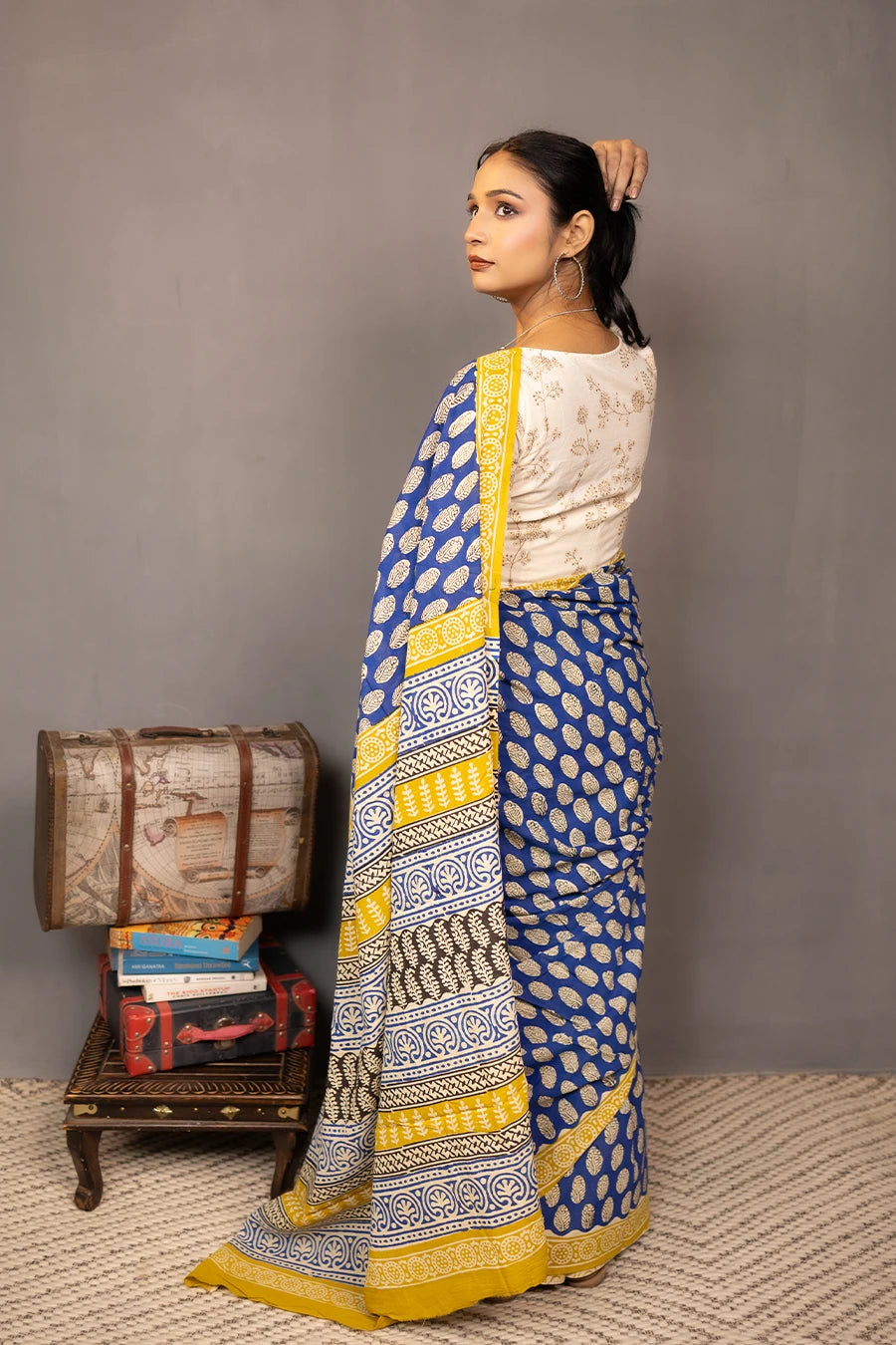 Woman wearing a blue and yellow saree standing next to vintage suitcases and books against a grey background
