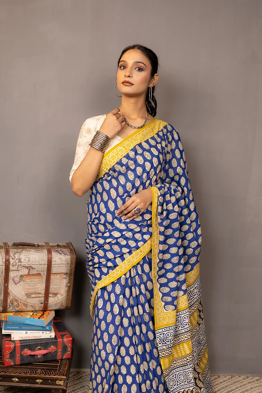 Woman wearing a blue and yellow saree standing next to vintage suitcases and books against a grey background